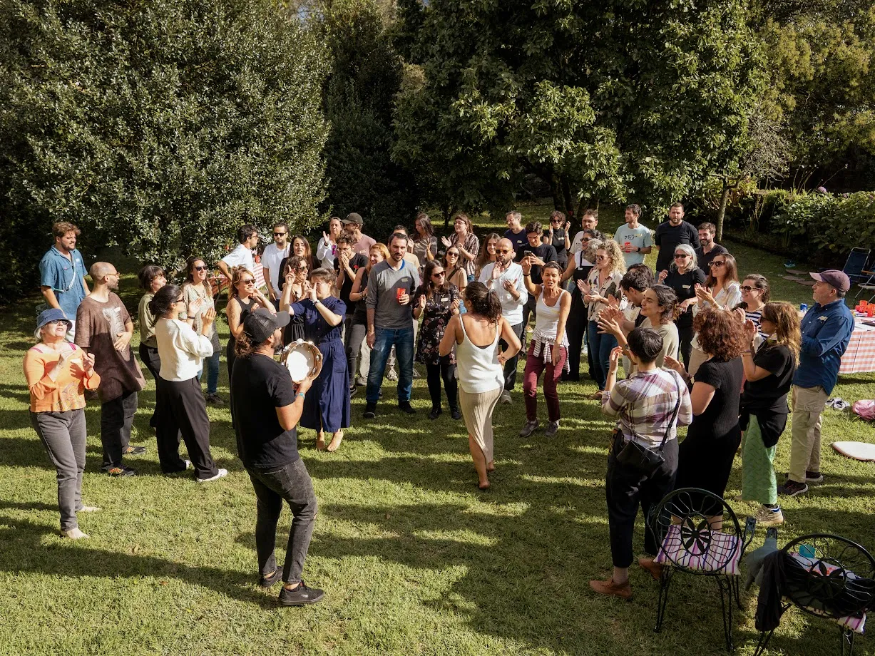 Crowd standing in a circle on a lawn clapping while a man plays a tambourine and a woman dances