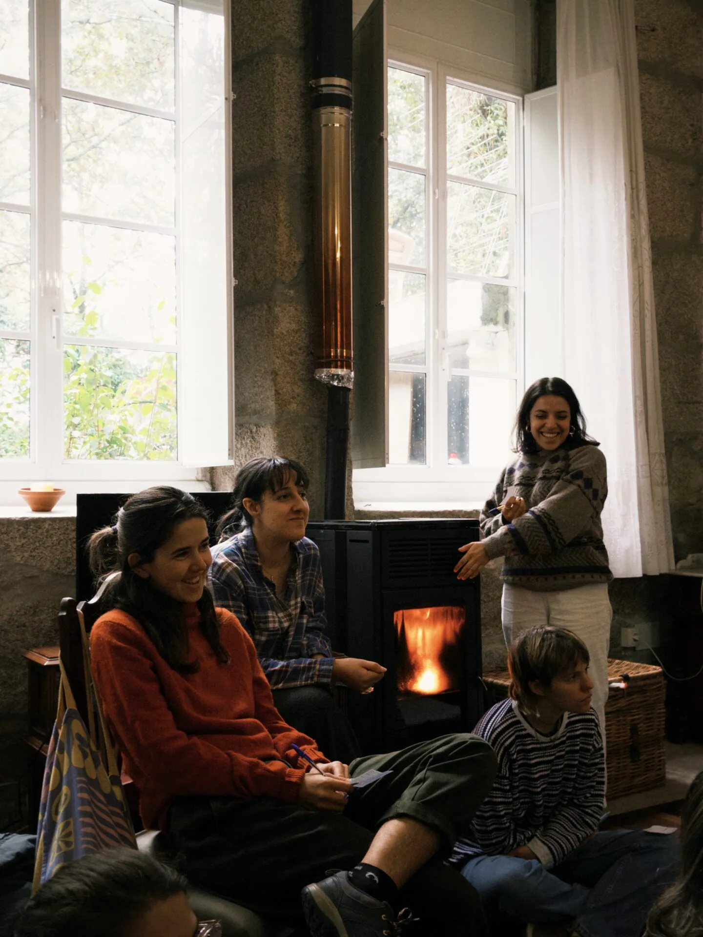 Two women and a child sitting together by a lit fireplace inside Anceu Coliving during the winter season