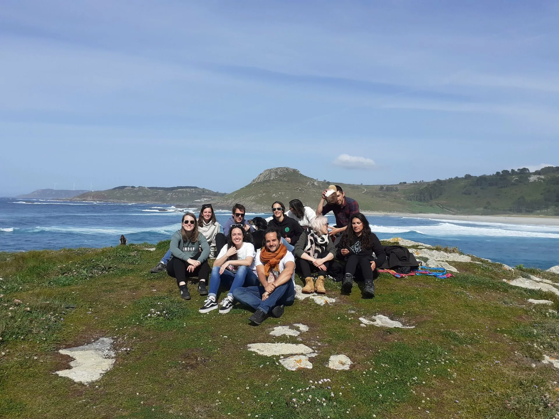 Eight people sitting close together on a grassy clifftop above a sandy beach and the sea