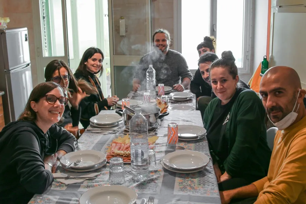 Nine people around a long dining table in a bright kitchen, steam rising from a pot in the middle