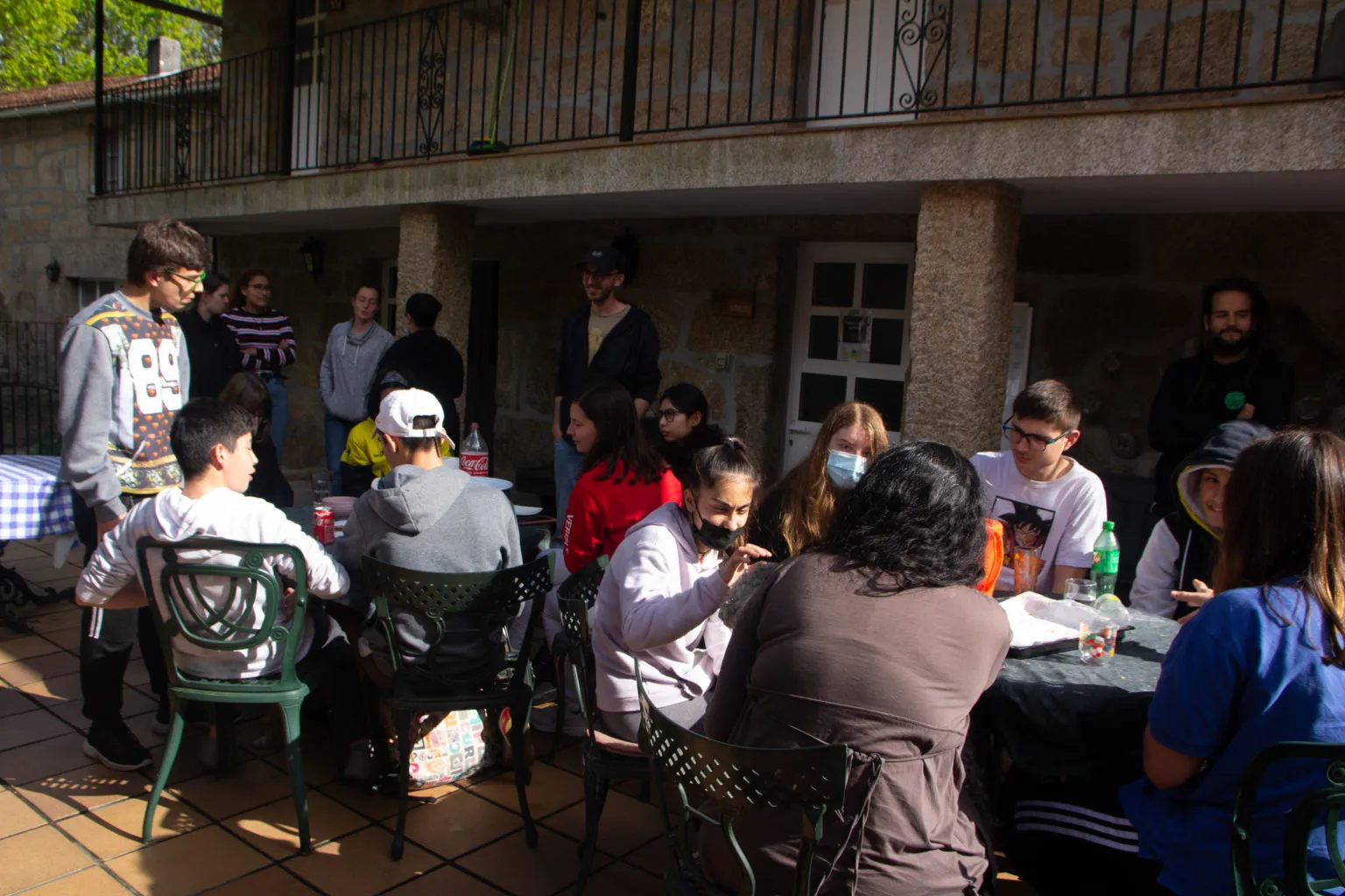 Young people and adults sitting at tables on a sunny stone patio under a balcony, some wearing face masks
