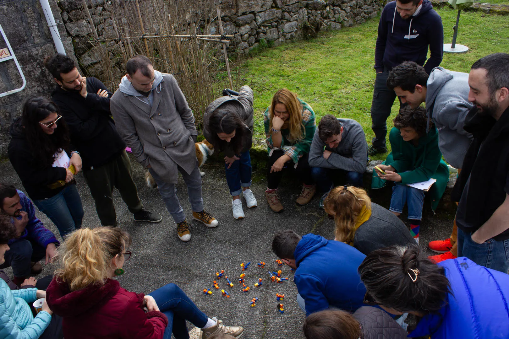 Seen from above, a large circle of people standing and crouching around small Lego models on a concrete yard