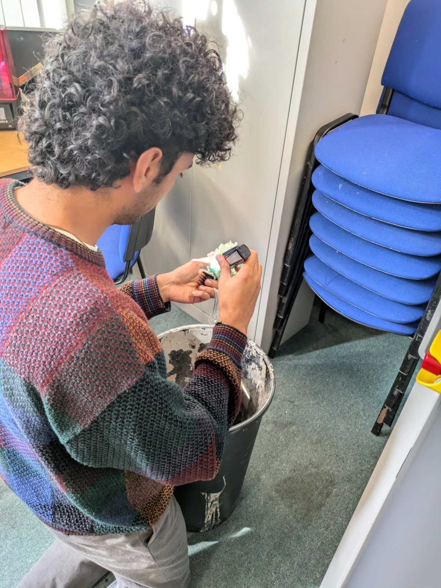 Man in a patterned jumper examining a small device and a green-flecked plastic piece over a bin in a workshop
