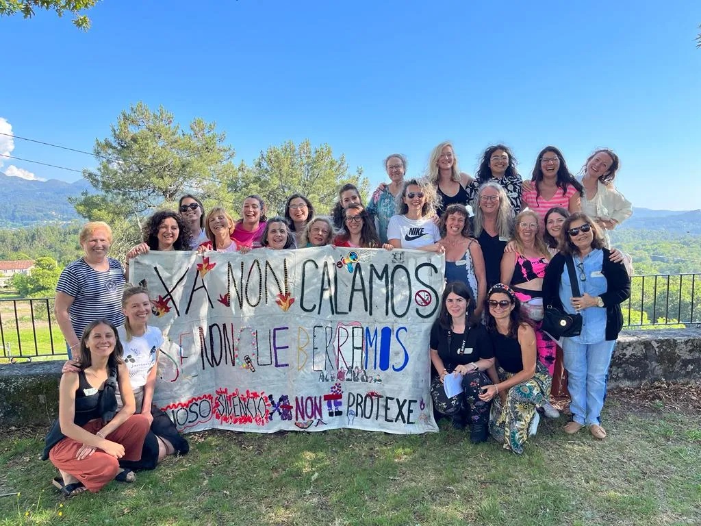 About twenty women holding a hand-painted banner reading “Xa non calamos, senón que berramos. O noso silencio non te protexe”