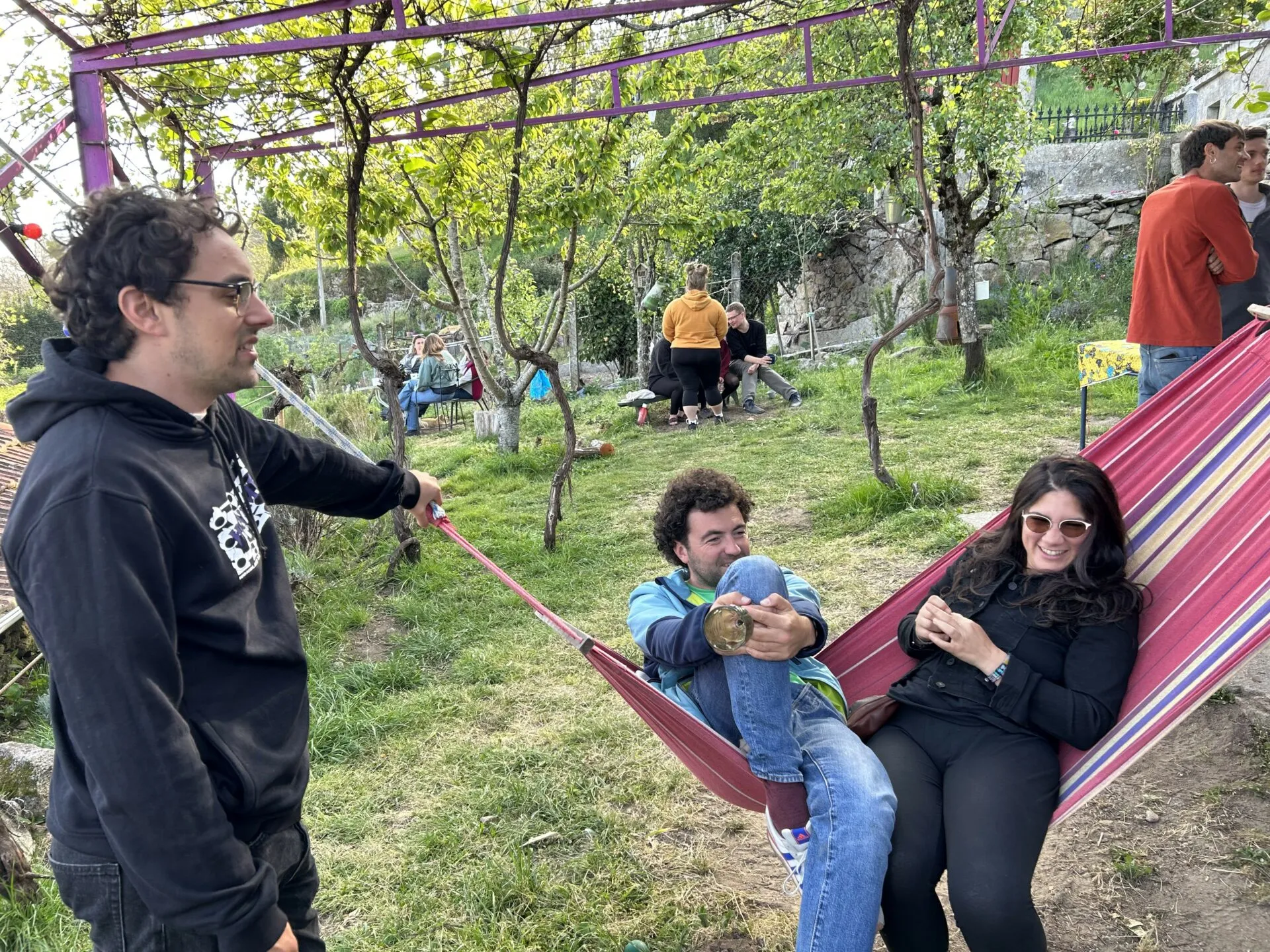 Two people sitting in a striped hammock under a pergola in a sunny garden while a man in a hoodie holds the hammock