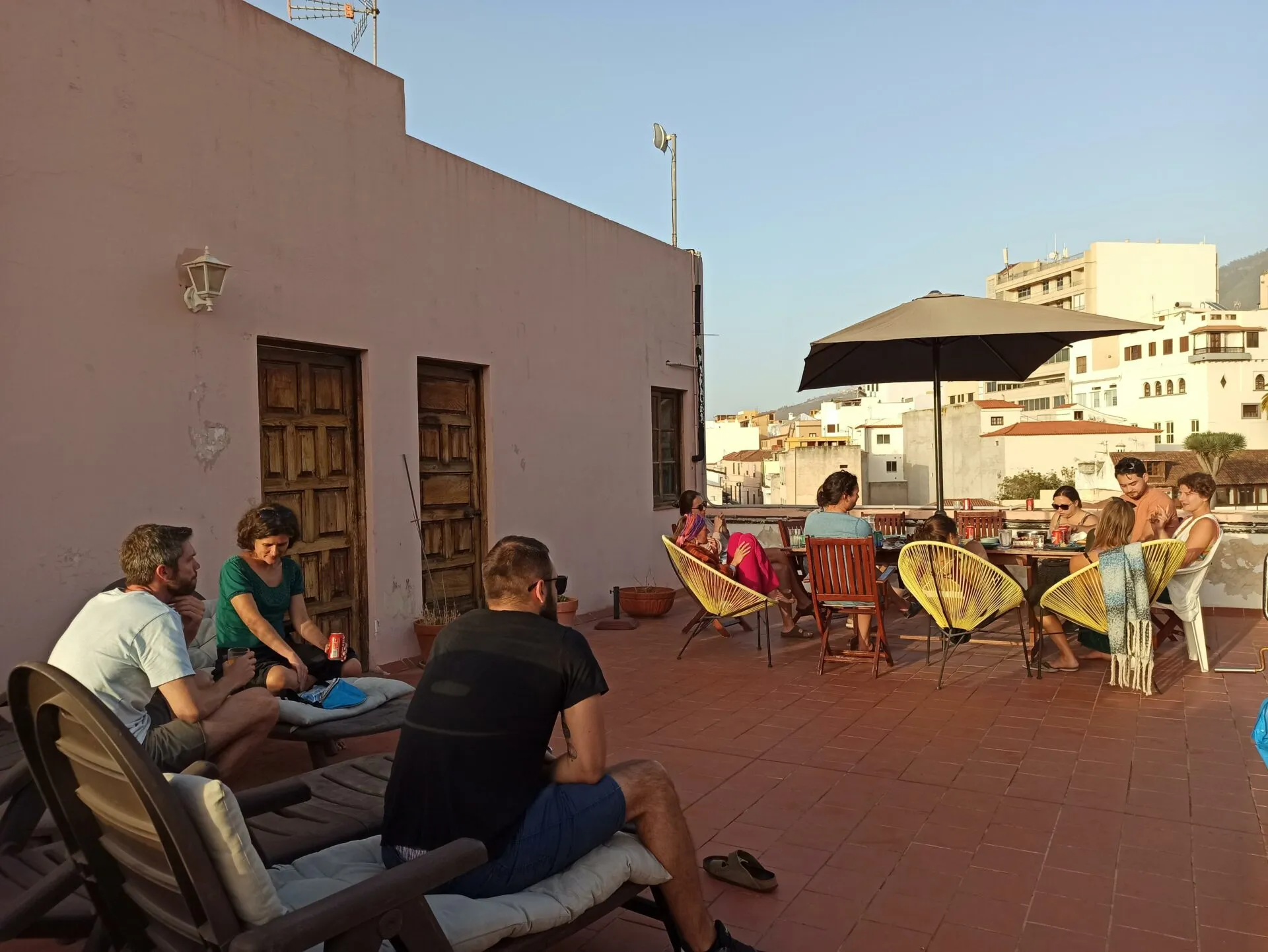 People relaxing at sunset on a rooftop terrace with yellow Acapulco chairs and a parasol, town rooftops behind