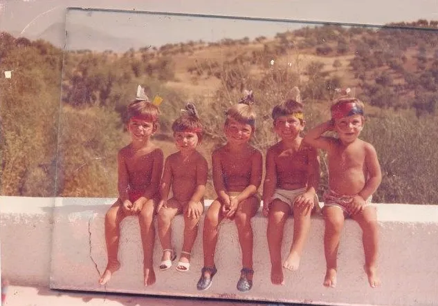 Faded old photograph of five young children in swimwear sitting in a row on a low white wall, dry hills behind
