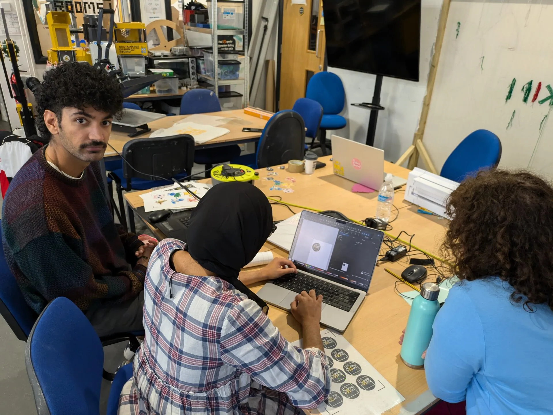 Three people working with laptops and sticker sheets at a cluttered makerspace bench