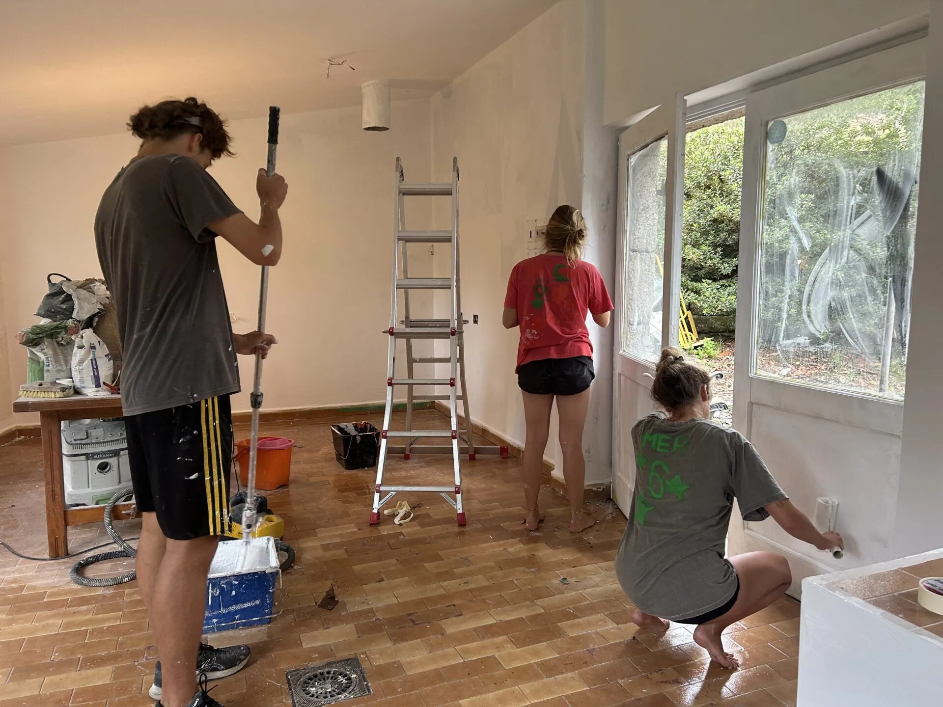 Three people sanding and painting the walls of an empty room, with a stepladder, paint buckets and a terracotta tiled floor