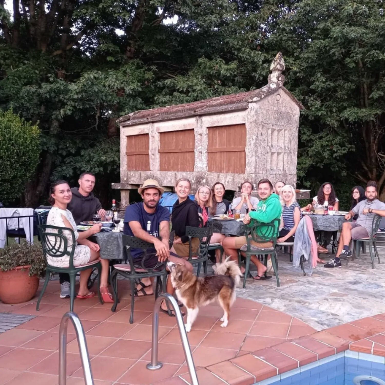 Around fifteen people having dinner at outdoor tables beside a pool, in front of a stone hórreo