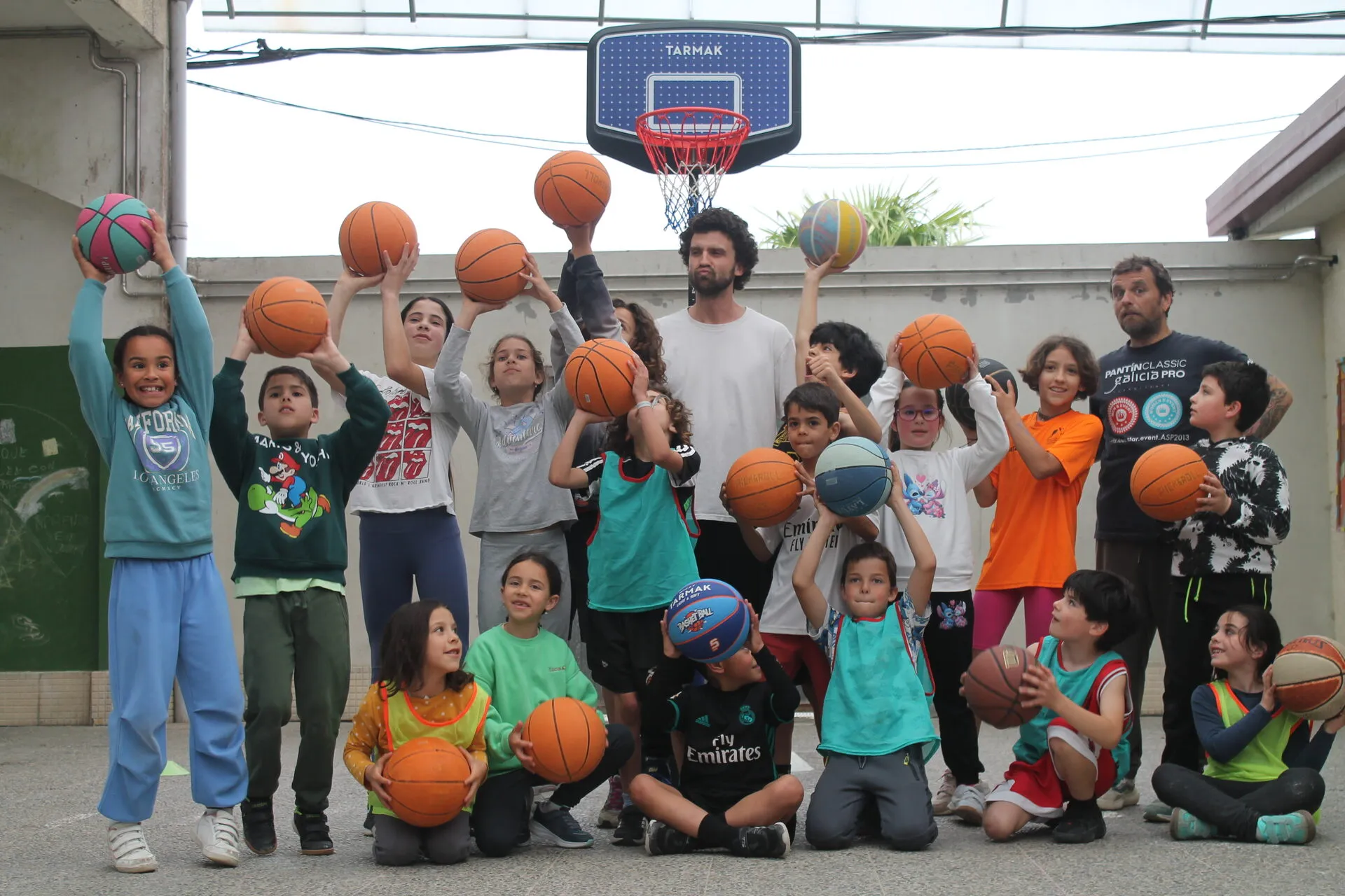 About twenty children holding basketballs up in the air under a hoop in a school yard, with Victor and another coach standing behind them