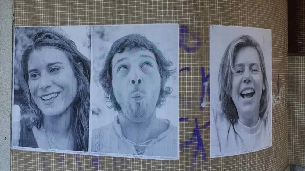 Three large black-and-white portrait posters of laughing faces pasted on a tiled wall covered in purple graffiti