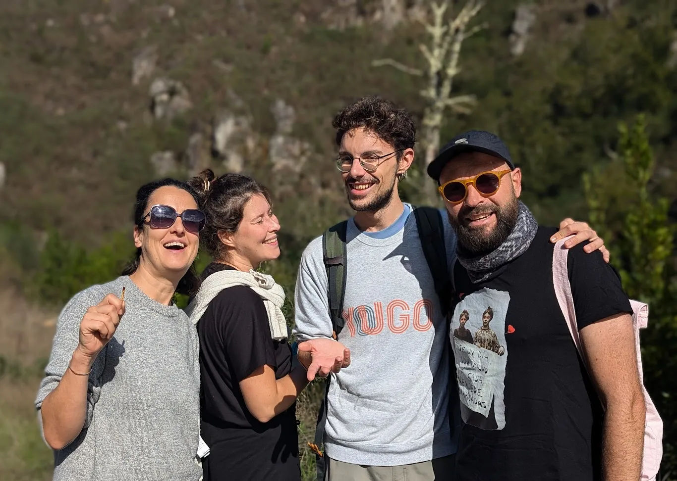 Four people laughing arm in arm outdoors, with a rocky scrub-covered hillside behind them