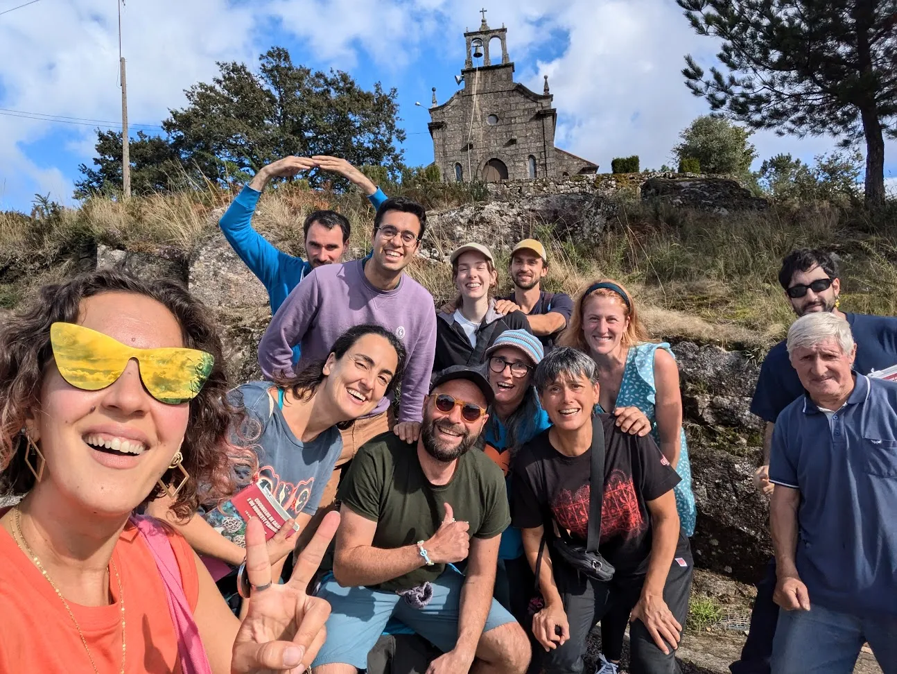 Group selfie of about a dozen people on a rocky hillside below a small stone chapel with a bell gable