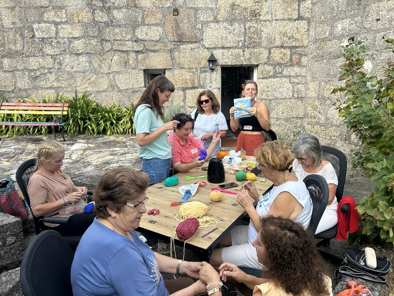 Women knitting and crocheting around a wooden table of coloured wool balls, against the granite wall of the house