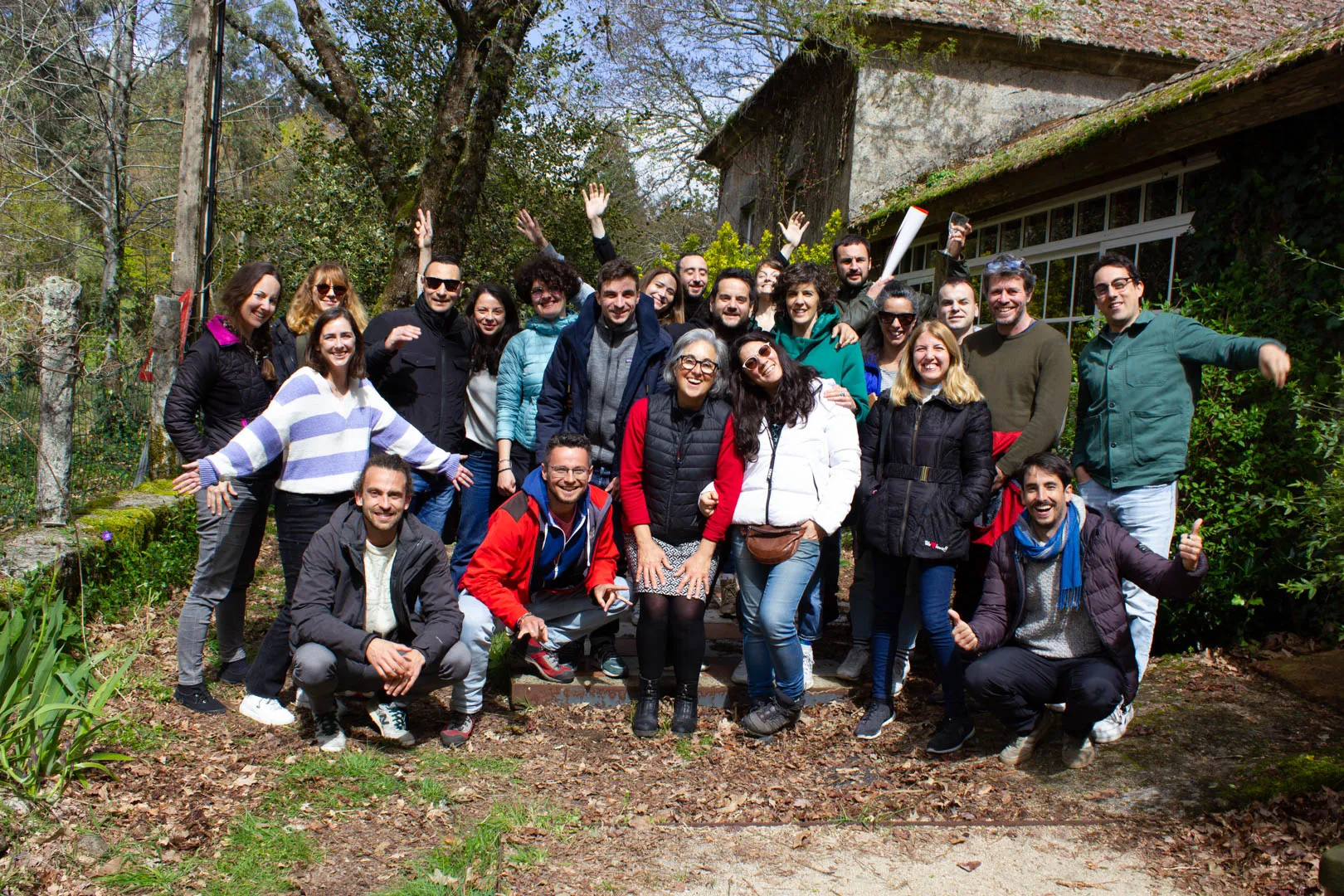 Around forty people posing in a leafy garden beside an old stone building, some with arms raised