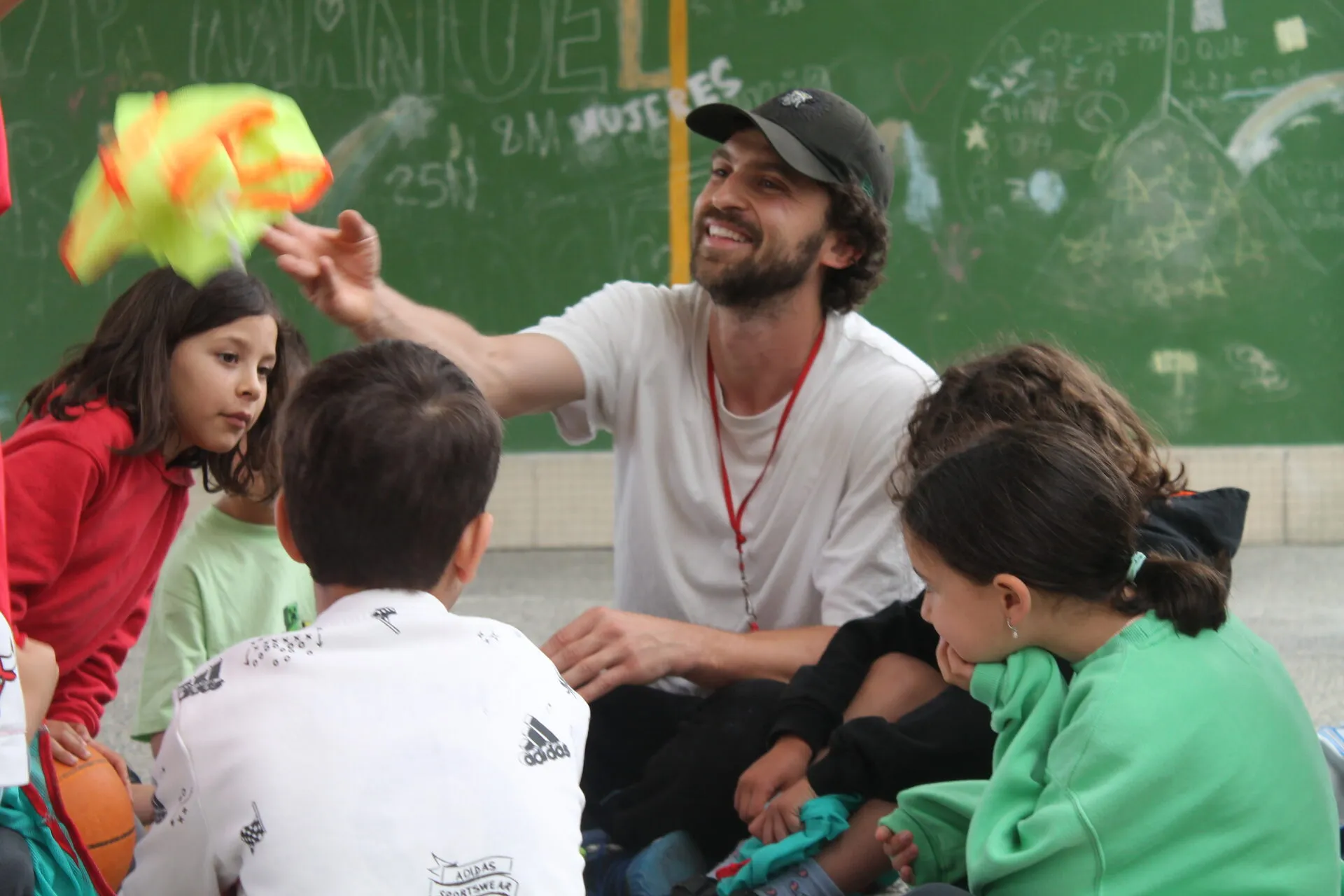 Victor sitting on the ground in a school yard with a group of children around him, holding up a bundle of coloured training bibs