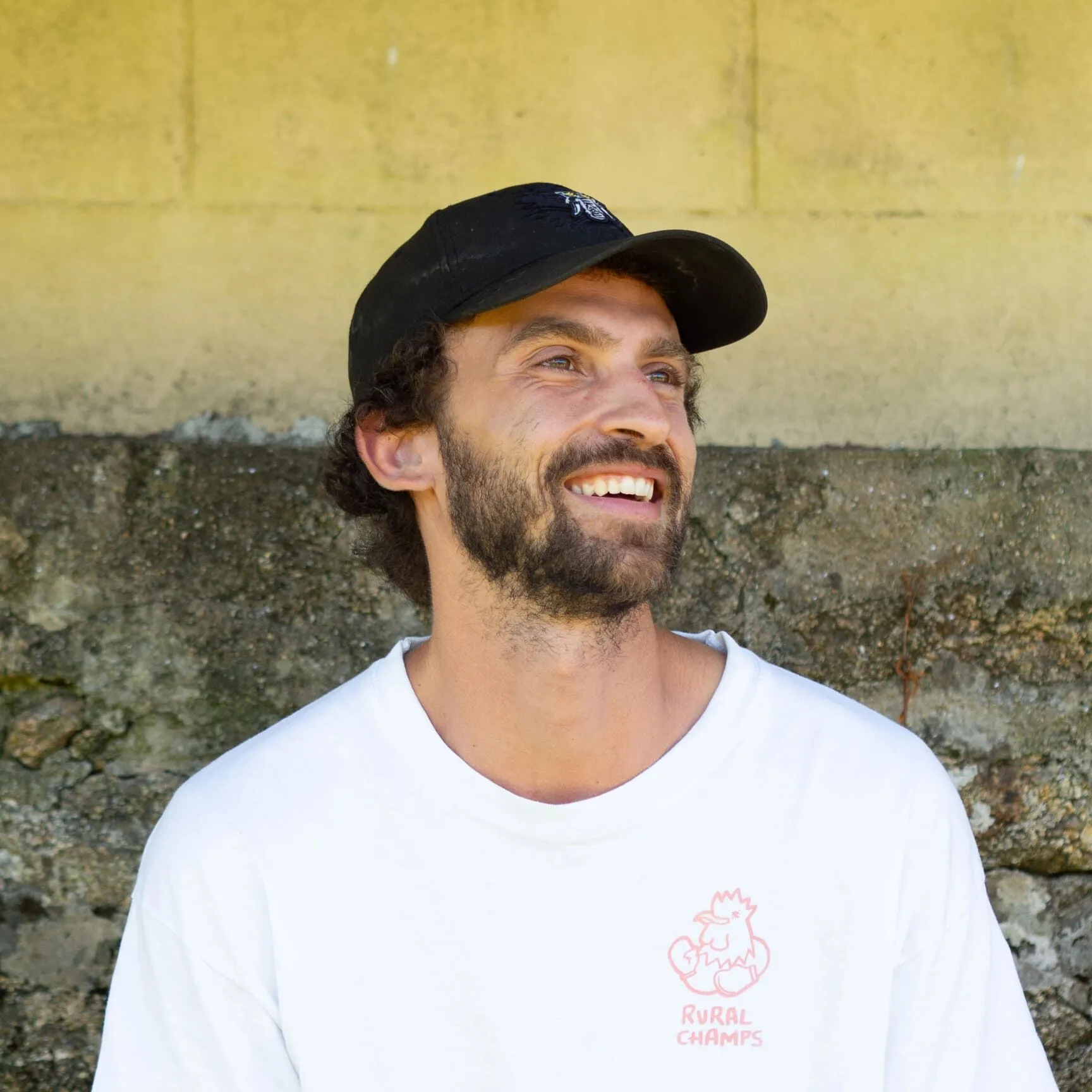 Victor smiling in a black cap and a white Rural Champs t-shirt, leaning against a yellow stone wall