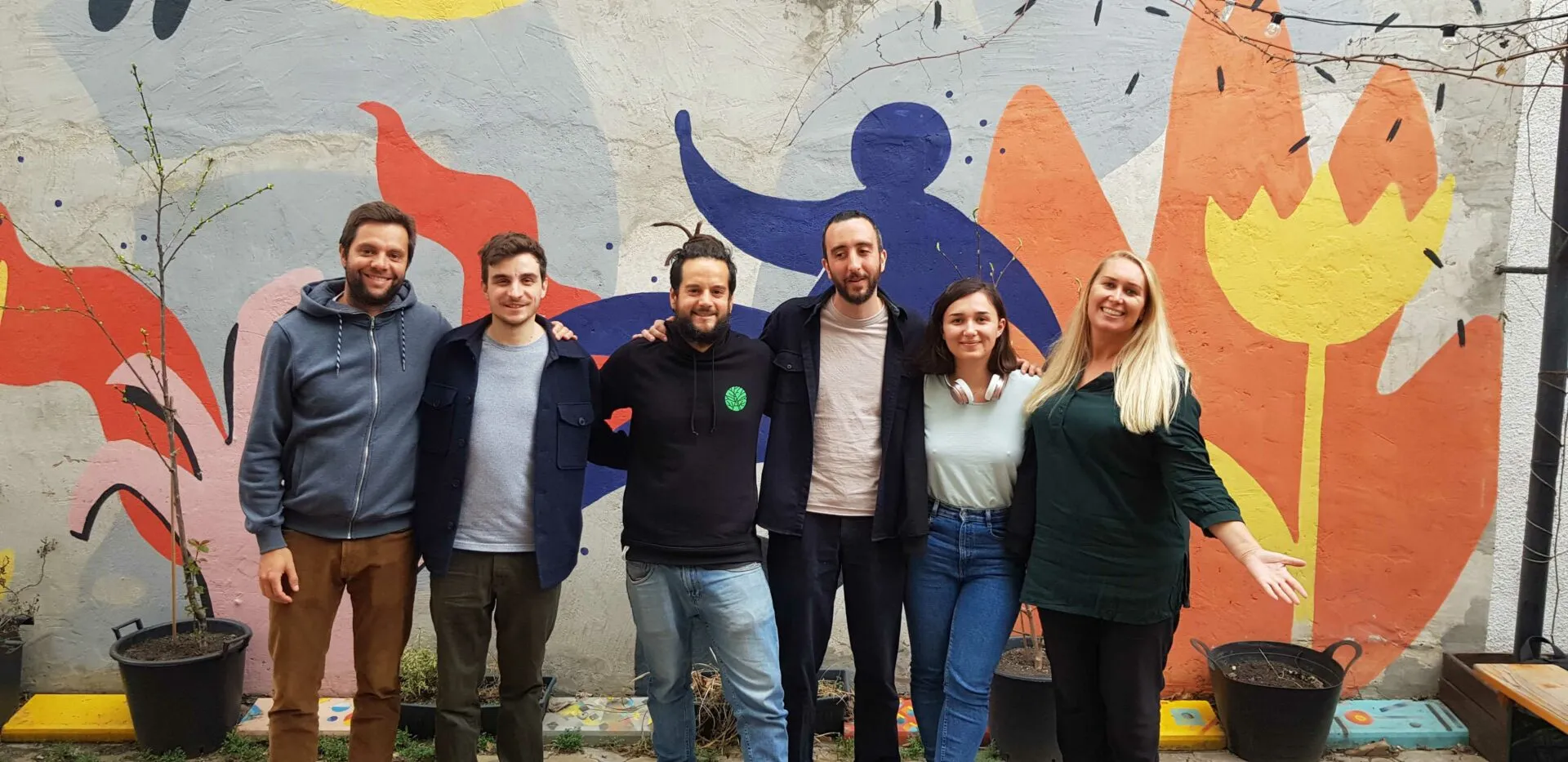 Six people arm in arm in a courtyard in front of a large colourful mural of a blue figure and flowers
